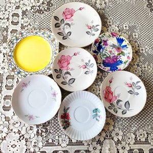 Può includere: Una collezione di sette tazze da tè e piattini vintage con motivi floreali. I piattini sono bianchi con bordo dorato e presentano vari disegni floreali, tra cui rose, foglie e altri elementi botanici.