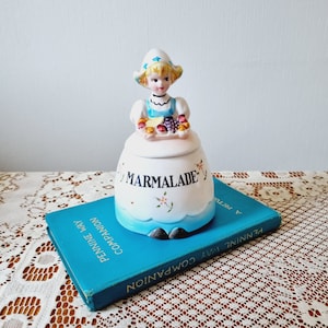 Peut inclure: Un pot à confiture en céramique vintage avec une figurine de fille sur le dessus. Le pot est décoré de fleurs bleues et blanches et du mot "MARMALADE".