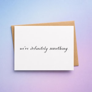 May include: A white greeting card with the text "we're definitely something" in black script. The card is on a brown envelope, set against a gradient background of blue and purple.