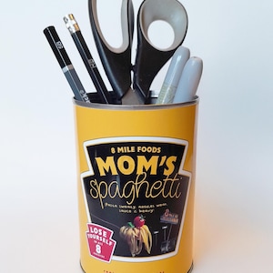 May include: A yellow can repurposed as a desk organizer, featuring the text "MOM'S spaghetti" and other text. The can holds pencils, pens, and scissors. The can's design includes a spaghetti dish illustration and the phrase "Lose Yourself in All 8 Varieties."