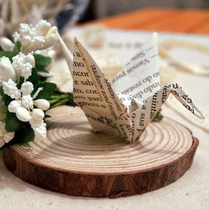 Pode incluir: Um grou de papel dobrado a partir de páginas de texto impresso assenta numa fatia circular de madeira. Ao lado do grou encontra-se um pequeno ramo de flores artificiais brancas com folhas verdes. O texto impresso é claramente visível.