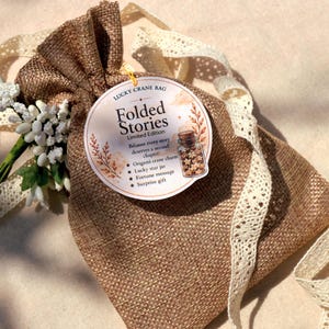 Pode incluir: Uma bolsa de serapilheira castanha com cordão, adornada com uma fita de renda cor de creme e um pequeno ramo de flores brancas. Uma etiqueta circular presa à bolsa apresenta o texto Folded Stories, Lucky Crane Bag e uma lista de itens.