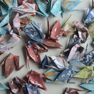 以下が含まれることがあります： 模様のある紙で作ったカラフルな折り鶴の集まり。鶴は様々な位置に折り畳まれ、薄い灰色の表面に散らばっている。