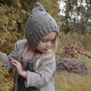 Peut inclure: Un bonnet en tricot marron clair avec un sommet pointu et de petits détails en forme de pompons sur la surface. Le bonnet présente un bord côtelé autour de l'ouverture du visage et est fabriqué dans un fil texturé, assurant un ajustement serré.