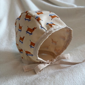 May include: A beige baby bonnet featuring a repeating pattern of brown dachshunds on blue skateboards. The bonnet has a light beige trim and a matching tie closure. The interior of the bonnet is a solid beige colour.