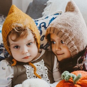 May include: Two children wearing knitted pixie hats. One hat is mustard yellow, the other is tan. The children are wearing olive green and white tops. A knitted pumpkin is in the foreground. The background includes a blue and white patterned cushion.