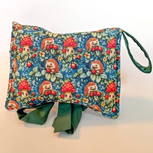 Peut inclure: Une pochette rectangulaire avec une poignée et des liens verts. Le tissu présente un motif fantaisiste d'hérissons, de champignons rouges et de baies sur fond bleu. La pochette est conçue pour transporter des objets.