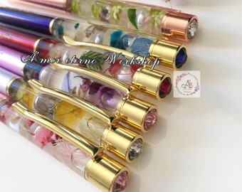 May include: A collection of handcrafted pens with clear barrels filled with dried flowers. The pens have gold-colored accents and gemstone-like tips in various colors. The text "Amor eterno Workshop" is visible.