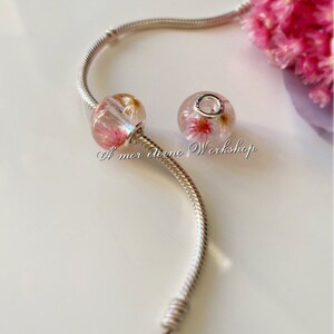 May include: Two clear glass beads with pink and gold floral inclusions are displayed on a silver bracelet. The beads are round and oval-shaped. The text "A mor eterno Workshop" is visible.