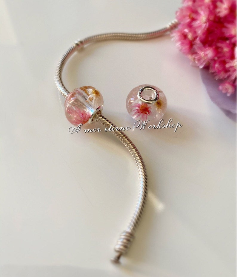 May include: Two clear glass beads with pink and gold floral inclusions are displayed on a silver bracelet. The beads are round and oval-shaped. The text "A mor eterno Workshop" is visible.