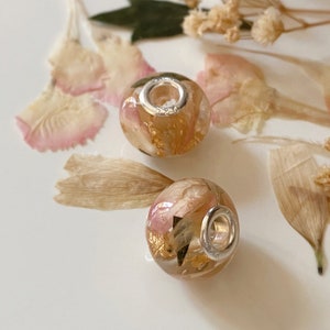 May include: Two round, clear beads with silver-colored metal rings. The beads are filled with dried pink and gold-colored floral elements. The beads are resting on a white surface, surrounded by dried flowers and petals.