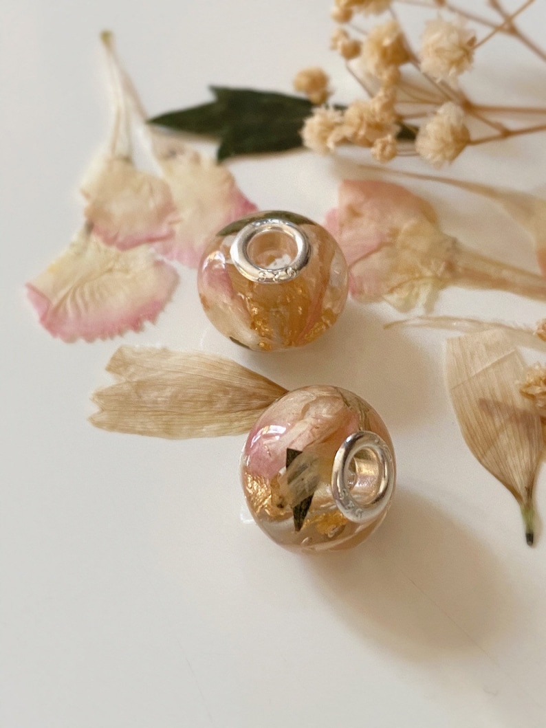 May include: Two round, clear beads with silver-colored metal rings. The beads are filled with dried pink and gold-colored floral elements. The beads are resting on a white surface, surrounded by dried flowers and petals.