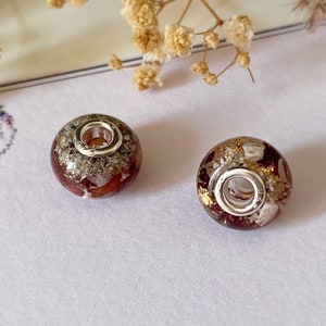 May include: Two round, decorative beads with a silver-colored core and a mix of red, gold, and white inclusions. The beads are set against a white background with dried flowers and a thank you card.
