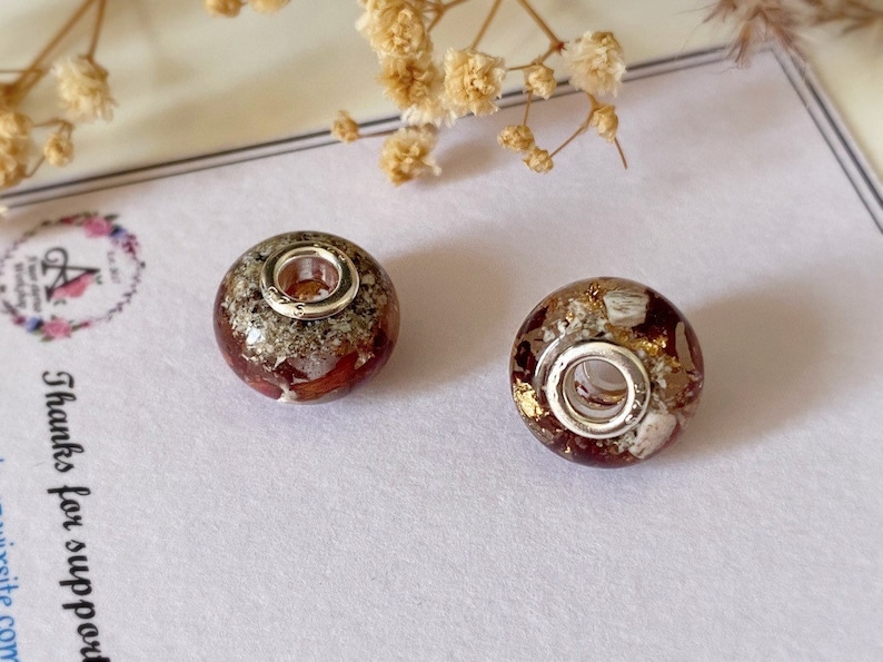 May include: Two round, decorative beads with a silver-colored core and a mix of red, gold, and white inclusions. The beads are set against a white background with dried flowers and a thank you card.