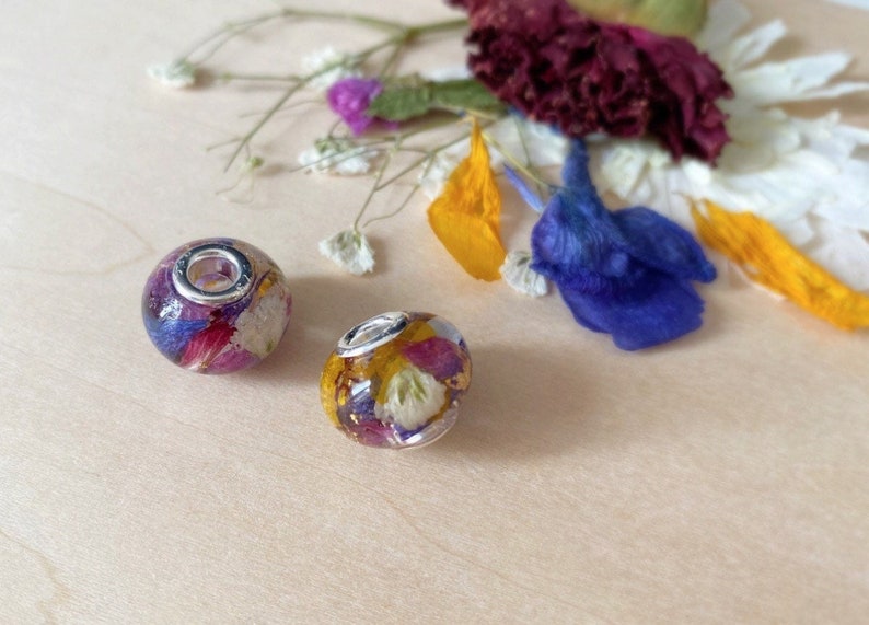 May include: Two clear, round beads filled with colorful dried flowers. The beads have a silver-colored metal core and are surrounded by purple, pink, yellow, and white petals. Dried flowers and petals are scattered around the beads.