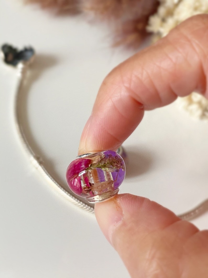 May include: A clear, round charm bead with a silver-colored metal base. The bead is filled with dried flowers in shades of pink, purple, and beige. The charm is held by a person's fingers, with a silver bracelet in the background.