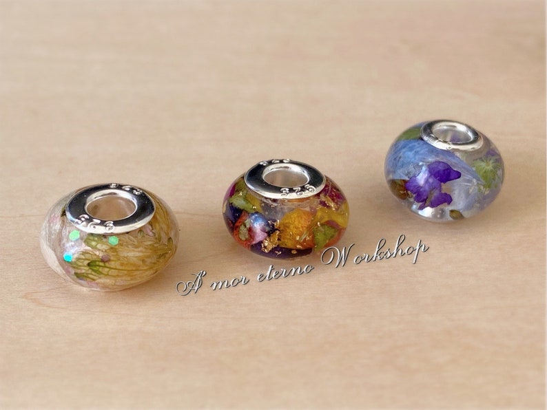 May include: Three glass beads with floral inclusions and silver-colored metal accents. The beads contain dried flowers in shades of yellow, purple, and green. The text "A mor eterno Workshop" is visible.