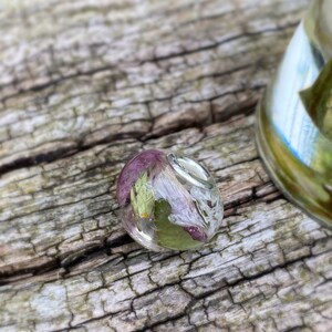May include: A clear, oval-shaped resin pendant with dried flowers and leaves encased inside. The flowers are a mix of purple and white, with green leaves. The pendant has a silver bail and is set against a weathered wood background.
