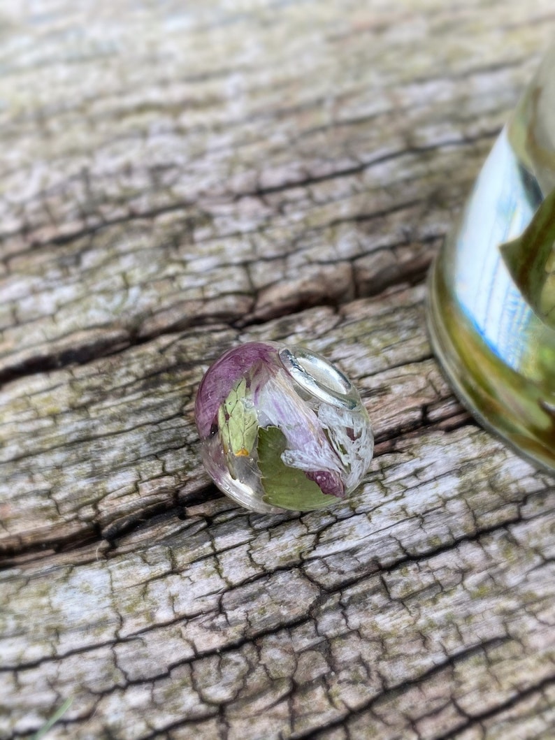 May include: A clear, oval-shaped resin pendant with dried flowers and leaves encased inside. The flowers are a mix of purple and white, with green leaves. The pendant has a silver bail and is set against a weathered wood background.