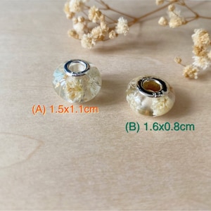 May include: Two clear resin beads with dried flowers and silver-tone metal accents. Bead A measures 1.5cm x 1.1cm, and bead B measures 1.6cm x 0.8cm. The beads are displayed on a light wood surface with dried flowers.