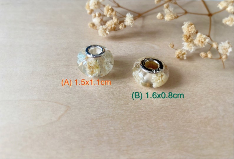 May include: Two clear resin beads with dried flowers and silver-tone metal accents. Bead A measures 1.5cm x 1.1cm, and bead B measures 1.6cm x 0.8cm. The beads are displayed on a light wood surface with dried flowers.