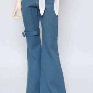 Op de afbeelding: Blauwe broek met uitlopende pijpen, voorzien van een zijband met een zilverkleurige gesp op het bovenbeen, gecombineerd met een wit topje met een striksluiting aan de voorzijde. De broek heeft een aansluitende taille en wijde pijpen.