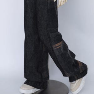 May include: Dark grey denim trousers with a wide leg design. The trousers feature a subtle plaid pattern in shades of brown and tan along the side seams. The trousers are paired with off-white trainers.