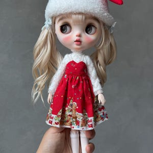 May include: A doll with blonde hair in pigtails wearing a red and white Santa hat, a white long-sleeved textured top, and a red dress featuring a festive holiday print with teddy bears and gifts. The doll wears white knee-high socks and black shoes.