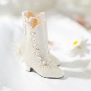 Op de afbeelding: Off-white enkellaarsjes met een kleine hak, voorzien van parelaccenten en een decoratief kanten ontwerp. De laarzen hebben een rits en staan tegen een zachte, bloemrijke achtergrond. De tekst "A OFF WHITE" staat onderaan.