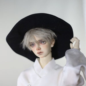 Pode incluir: Uma boneca com cabelo prateado usando um chapéu preto de aba larga e uma camisa branca de colarinho com dois botões prateados.