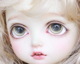 May include: Close-up of a porcelain doll with blonde curly hair and large green eyes. The doll has a delicate facial features and a soft, pink blush.