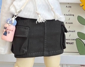 May include: A doll wearing a white sweater with a black letter 'M' on the front, a black skirt with pockets, white socks, and gray sneakers. The doll has brown hair in braids and is holding a small pink bottle.