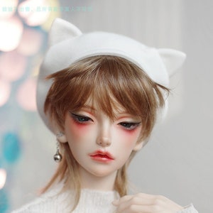 May include: A doll with brown hair wearing a white knitted hat with cat ears and a white sweater. The doll has green eyes and red blush on its cheeks. The sweater has a small blue logo with the text "喵猫 MiuCat"