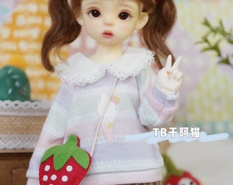 May include: A doll with brown hair in pigtails wearing a light purple and pink striped sweater with a white collar, a brown plaid skirt, white socks, and brown shoes. The doll is holding a red felt strawberry purse with white polka dots and green leaves.