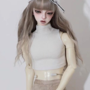 May include: A doll with long, wavy, light brown hair wearing a white crop top, tan trousers, and a white baseball cap. The doll has a pale complexion and is posed against a white background. The doll's outfit is casual and modern.