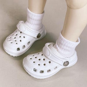 May include: A pair of white clogs with multiple ventilation holes, worn with white socks. The clogs have a textured surface and a pivoting heel strap. The clogs are made of a lightweight material and feature a silver-colored button on the side.
