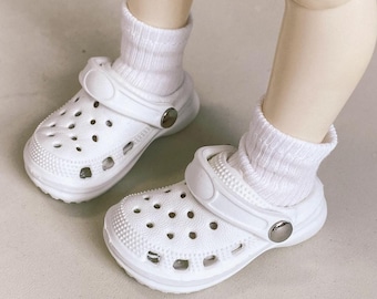 May include: A pair of white clogs with multiple ventilation holes, worn with white socks. The clogs have a textured surface and a pivoting heel strap. The clogs are made of a lightweight material and feature a silver-colored button on the side.