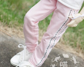 Peut inclure: Pantalon de survêtement rose avec des bandes latérales noires qui disent "Meow" à plusieurs reprises. Le pantalon de survêtement est porté par une poupée avec des baskets blanches. La poupée tient un skateboard jouet bleu et blanc.