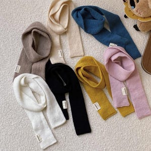 May include: A collection of ten colourful knit scarves in various colours, including beige, brown, blue, black, white, pink, and yellow. The scarves are arranged in a semi-circle on a light beige surface.