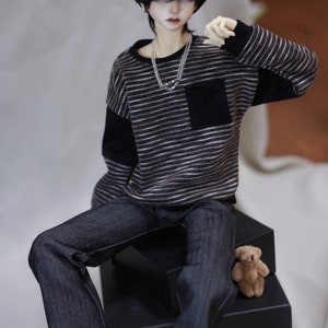 May include: A doll wearing a black and white striped sweater with a black pocket, black jeans with rolled up cuffs, and a black beret. The doll is sitting on a black box with a small brown teddy bear on top.