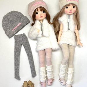 May include: Two dolls wearing white fuzzy jackets, hats with "Unisamon" text, and matching outfits. Accessories include gray leggings, shoes, and a gray beanie. The dolls are styled with pink and beige accents.