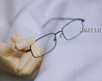 May include: Black rectangular glasses with a thin metal frame. The glasses are held in a hand and the text "SNAKE EYES" is visible in the background.