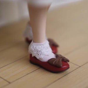 Könnte beinhalten: Ein Paar rote Schuhe mit einer braunen Schleife vorne. Die Schuhe werden von einer Puppe mit weißen Spitzensocken getragen.