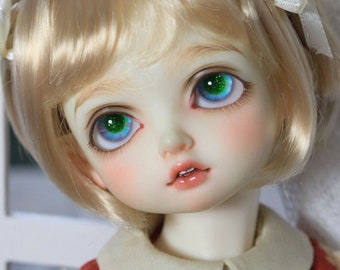 May include: A close-up of a doll's face with blonde hair and green eyes. The doll is wearing a white collar and a red dress.