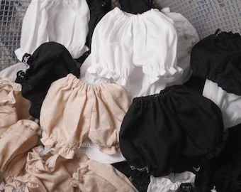 Op de afbeelding: Een verzameling poppenkleding in wit, zwart en beige. De poppenkleding omvat blouses met gerimpelde mouwen en bloomers. De stof lijkt licht en delicaat.