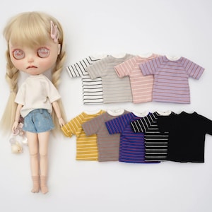 May include: A doll wearing a white t-shirt and denim shorts, with several miniature striped t-shirts in various colors. The shirts include black and white, gray, pink, and purple stripes, as well as yellow, brown, blue, and black.
