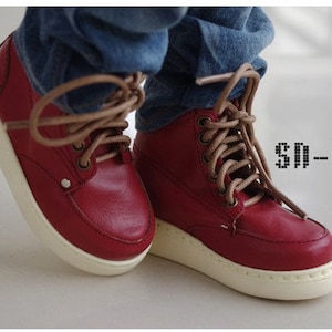 Op de afbeelding: Een paar rode leren hoge sneakers met bruine veters. De sneakers hebben een witte zool en worden gedragen door iemand die een blauwe jeans draagt.