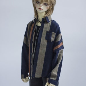 May include: A doll wearing a blue and brown striped long-sleeved shirt with a pocket on the left side. The doll is also wearing black shorts and has cat ears.