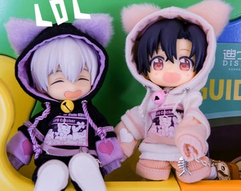 May include: Two small dolls wearing cat-themed hoodies. The doll on the left is wearing a black hoodie with purple ears and a pink heart on the sleeve. The doll on the right is wearing a pink hoodie with white ears and a pink heart on the sleeve. Both dolls have a pink and white graphic on the front of their hoodies that says "Cute Kitty Pro Catcher".