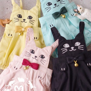 Puede incluir: Cuatro pares de petos con temática de gato en amarillo, azul, rosa y negro. Cada par presenta una cara de gato con una pajarita y una campana.
